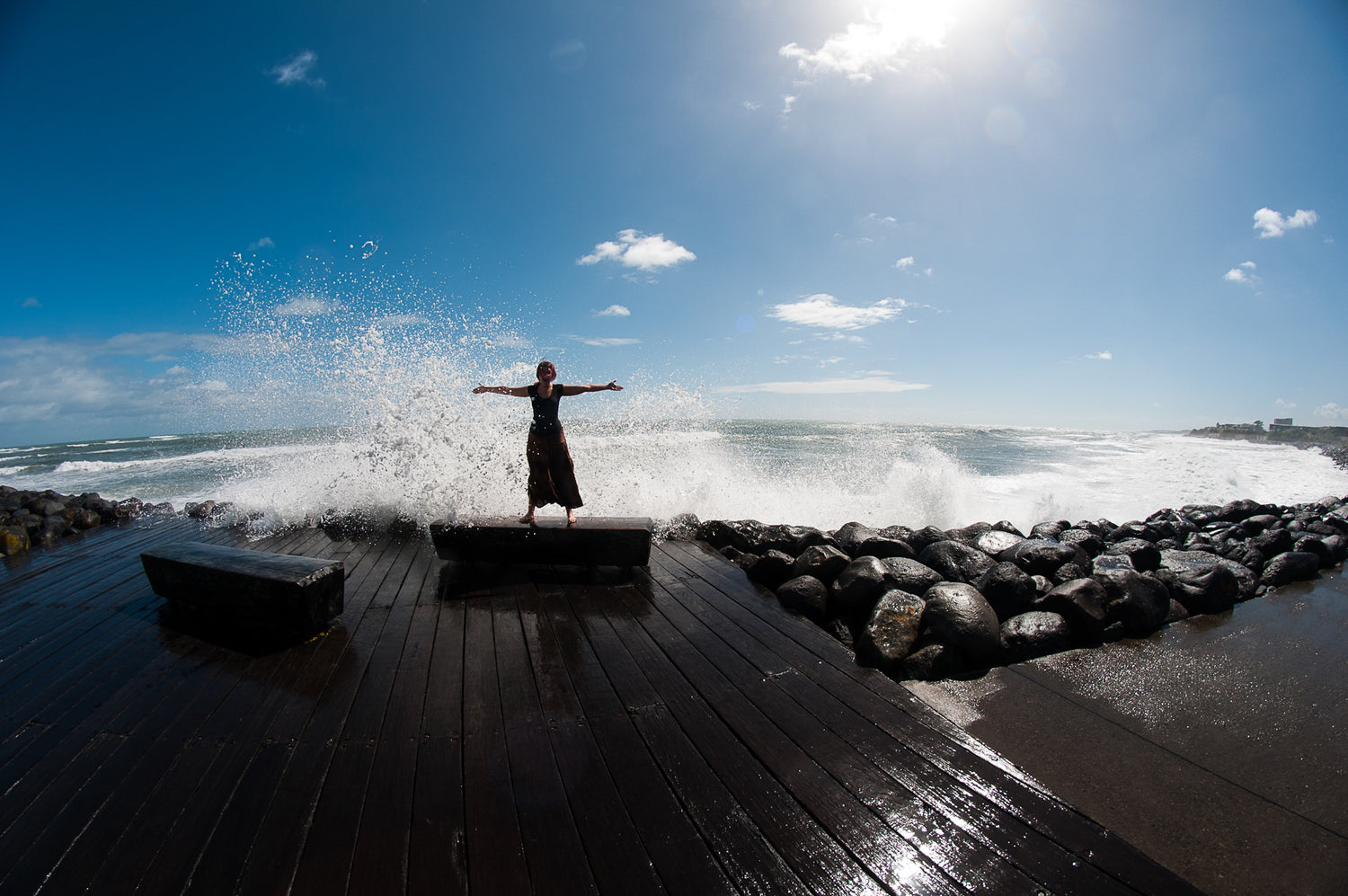 A woman in a skirt, photographer Shae Fischer, holds her arms open wide standing on a sea wall welcoming and incoming wave from the turbulent seas behind her.